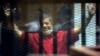 FILE - Former Egyptian President Mohamed Morsi, wearing a red jumpsuit that designates he has been sentenced to death, raises his hands inside a defendants cage in a makeshift courtroom at the national police academy, in an eastern suburb of Cairo.