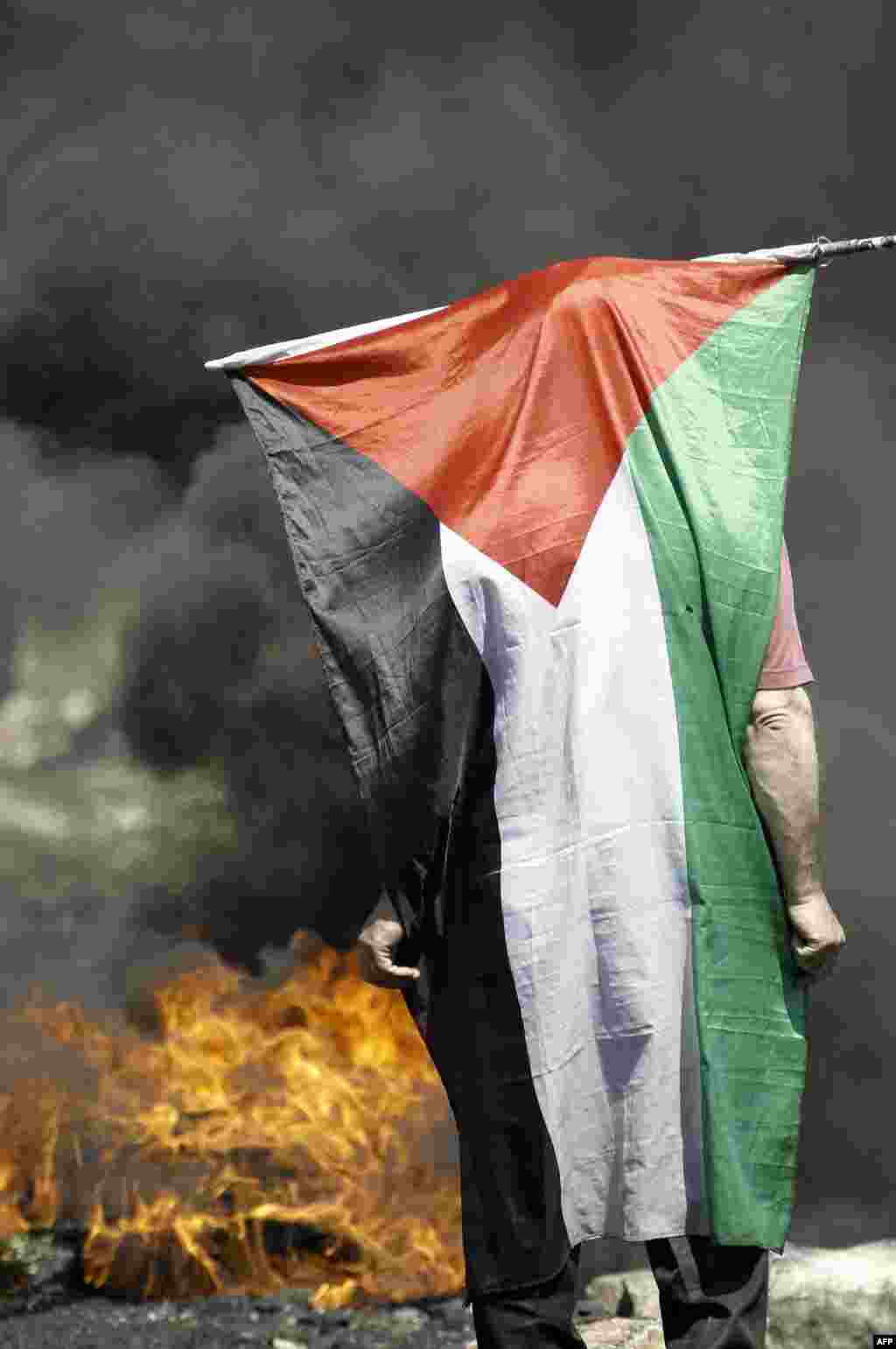 Seorang pengunjuk rasa warga Palestina berdiri dengan bendera negaranya tersampir di punggungnya di dekat ban-ban yang terbakar dalam bentrokan dengan tentara Israel setelah sebuah demonstrasi menentang pengambilalihan tanah Palestina oleh Israel di daerah pendudukan Tepi Barat. 